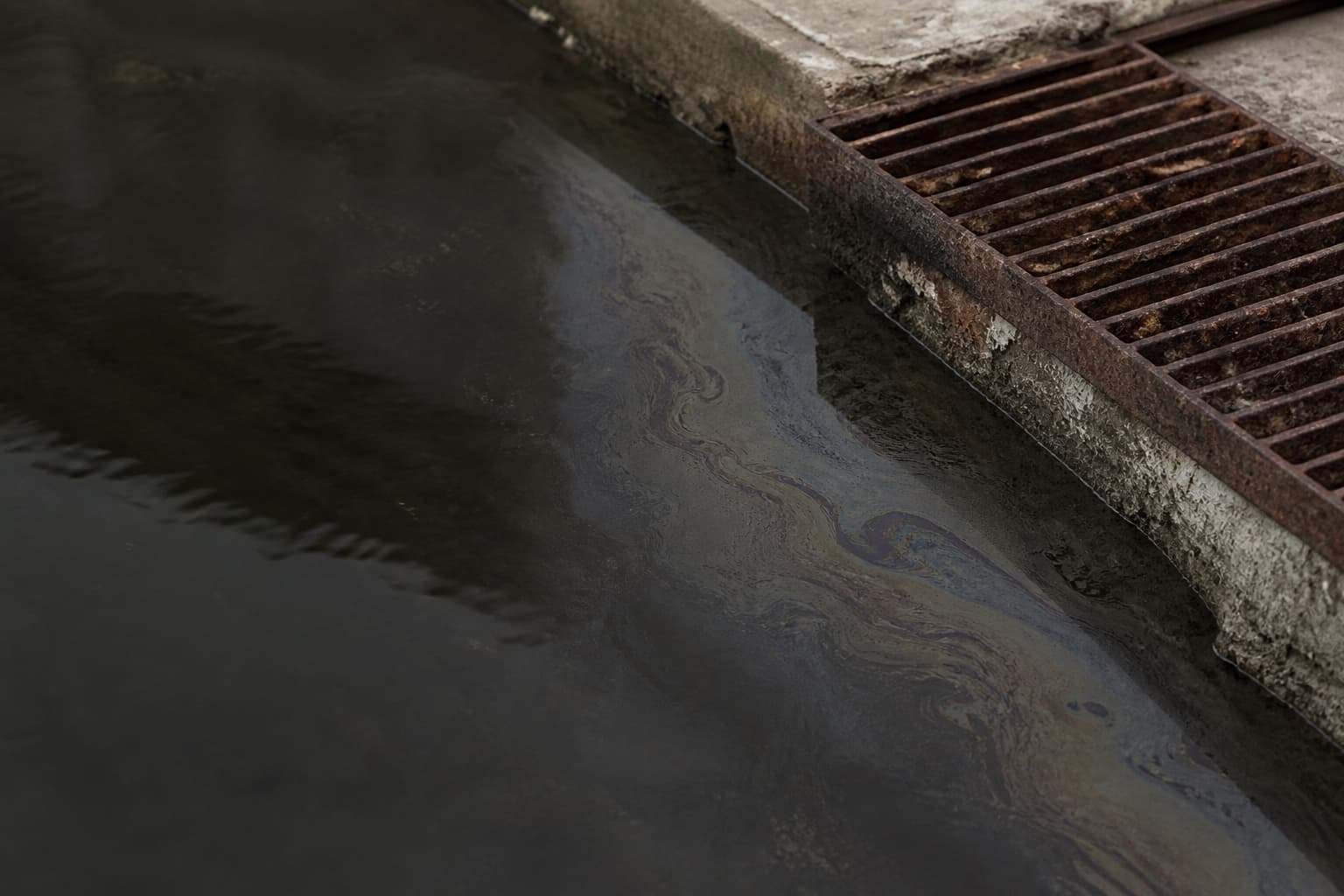 Película de hidrocarburo irisada sobre el agua detenida, junto a una rejilla de desagüe oxidada